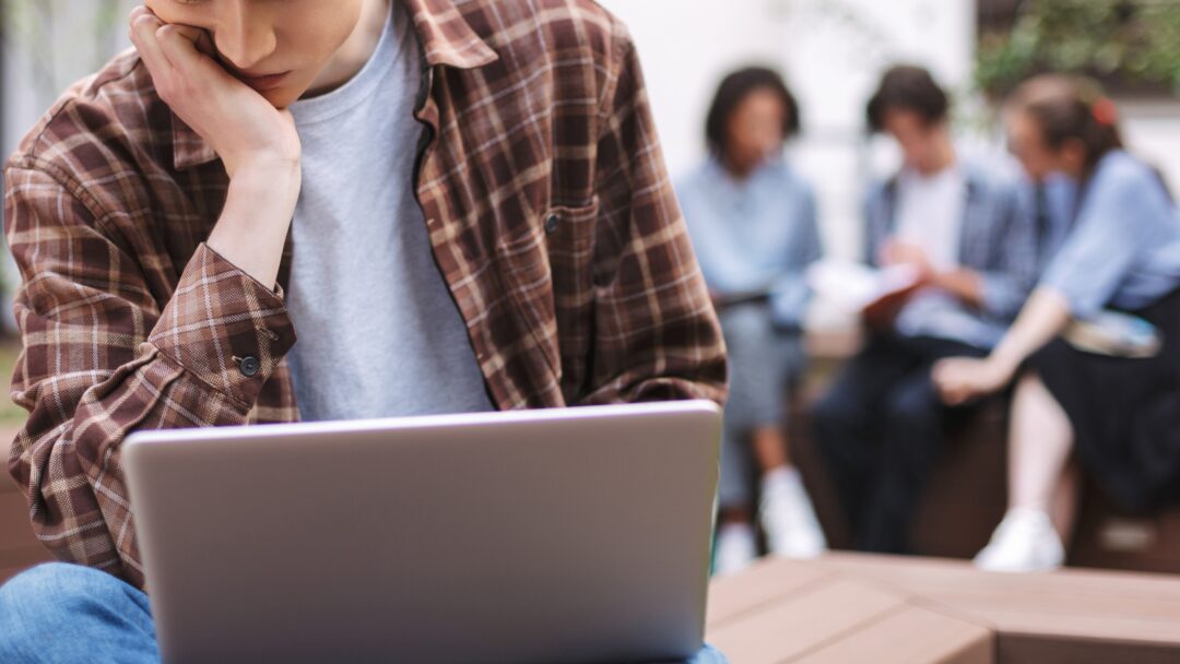 Photo of young tired man sitting on bench with laptop on knees and thinking while spending time in courtyard with students on background