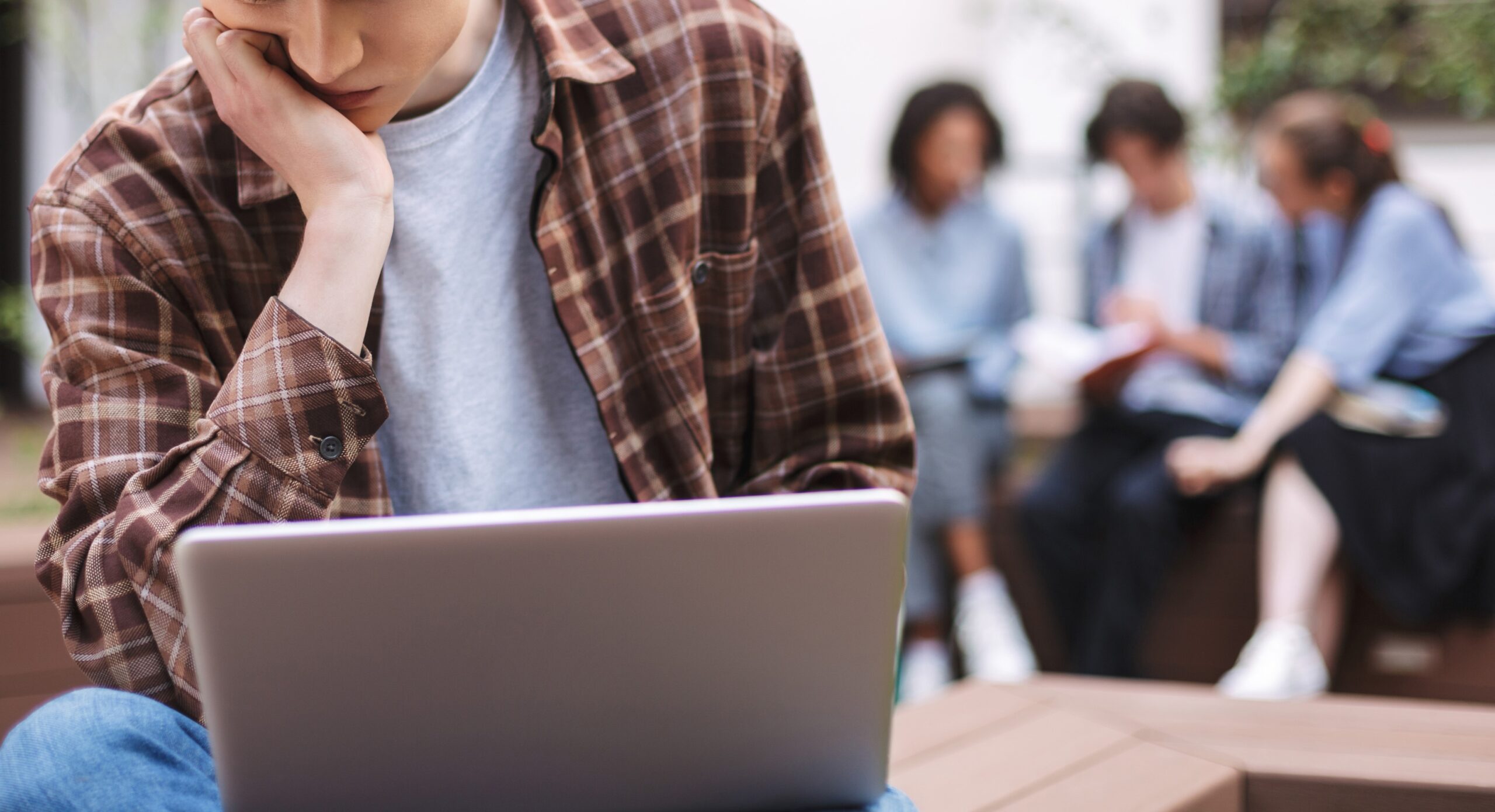 Photo of young tired man sitting on bench with laptop on knees and thinking while spending time in courtyard with students on background